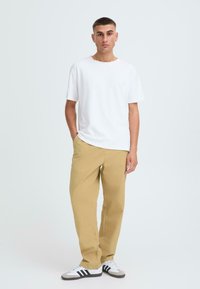 Weißes Baumwoll-T-Shirt, leicht khakifarbene Hose und schwarz-weiße Sneakers. Das Model steht lässig mit einer Hand in der Tasche vor einem einfarbigen Hintergrund.