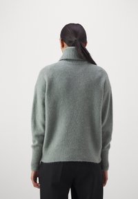 Grau, oversized Strickpullover mit hochgeschlossenem Kragen. Weicher Stoff, bequeme Passform und gerippte Bündchen und Saum. Rückansicht, getragen mit schwarzen Hosen.