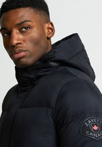 Melnā pūkainā jaka ar augstu apkakli un regulējamu kapuci. Kreisajā piedurknē ir uzlīme ar uzrakstu "ZAVETT" un sarkano kļavas lapu.