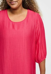 Fuchsia Bluse aus strukturiertem Stoff, mit Rundhalsausschnitt und drei Viertel Puffärmeln. Das Muster zeigt schwache senkrechte Linien.