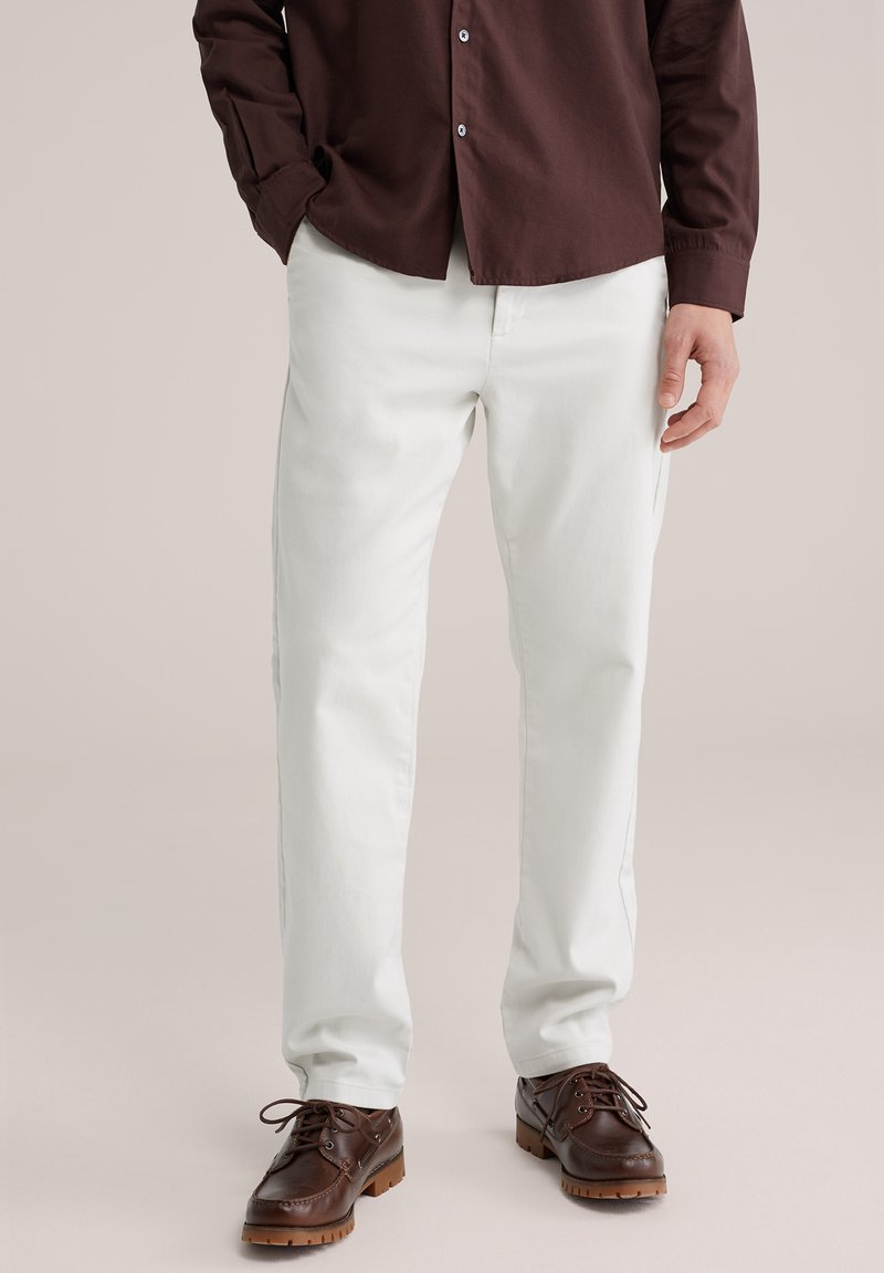 Uomo che indossa pantaloni bianchi aderenti, camicia marrone scuro con bottoni e scarpe di pelle marrone scuro, in piedi contro uno sfondo neutro.