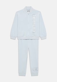Ensemble de survêtement bleu clair comprenant une veste zippée avec un logo "GUESS" en sequins et un pantalon assorti avec un détail brodé "G". Tissu doux.