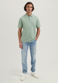 Lys grønn polo skjorte med krage, korte ermer og logo-detalj, sammen med lyseblå jeans og hvite joggesko.