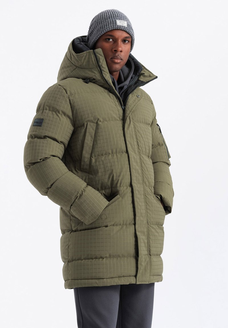 Chaqueta acolchada de color verde oliva con textura acolchada, capucha grande, bolsillos frontales y un parche negro. Llevada sobre un suéter gris.