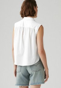 En person som bär en ärmlös vit skjorta och ljusblå denimshorts, sedd bakifrån mot en enkel ljus bakgrund.