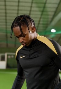 Fekete sportos felső arany Nike logóval és galléros díszítéssel, testhez simuló fazonban. Rövid fonatok és zöld beltéri fű háttér.