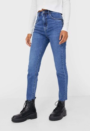 Persoon die een hoge taille blauwe skinny jeans met versleten zomen draagt en zwarte veterlaarsjes, gecombineerd met een wit langarmshirt.