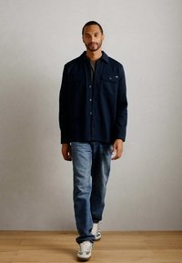 Man wearing a dark navy button-up jacket, gray shirt, blue jeans, and white sneakers walking on a light wooden floor against a gray wall.