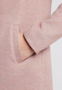 Ljusrosa texturerat tyg med en sidoficka och en något lös mudd. Detaljer inkluderar synliga sömmar och en mjuk yta.