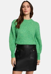 Maglione verde lavorato a maglia con maniche lunghe, scollo rotondo e texture a coste, abbinato a una mini gonna in pelle nera con tasche frontali.