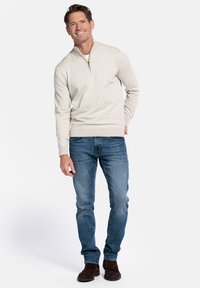 Beige sweater met ritsje en hoge hals, gemaakt van gestructureerde stof, gecombineerd met blauwe spijkerbroek en donkerbruine suède laarzen, gedragen door een staande man.