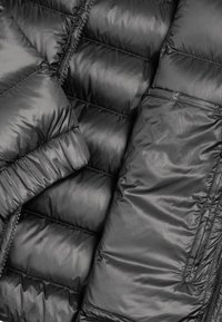 Giacca a vento nera con design trapuntato, texture liscia e tasca con zip. Il tessuto sembra lucido e resistente all'acqua.