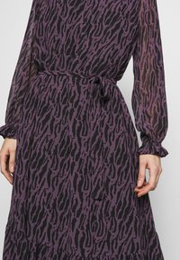 Robe violette et noire avec un motif rappelant l'imprimé animal, dotée de manches longues, d'une ceinture à nouer et de poignets à volants. Tissu léger et transparent.