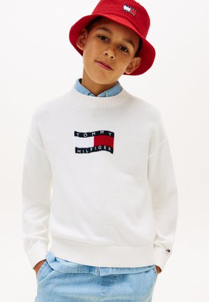 Dreng iført hvid Tommy Hilfiger sweater, rød bøllehat og lyseblå jeans, stående med hænderne i lommerne mod en ensfarvet baggrund.