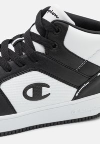 Sneaker alta in bianco e nero realizzata in pelle sintetica, con un pannello testurizzato, lacci neri e un logo del marchio ben visibile sul lato.