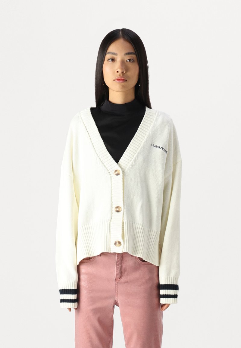 Cardigan crema con scollatura a V, cinque grandi bottoni e orlo a costine. Presenta accenti a righe scure sulle maniche. Abbinato a pantaloni rosa.