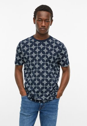 Man met kort krullend haar, gekleed in een marineblauw T-shirt met witte geometrische patronen en een blauwe spijkerbroek, staand met handen in de zakken.