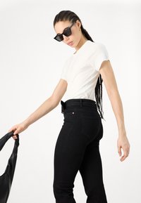 Valkoinen slim-fit t-paita yhdistettynä korkeavyötäröisiin mustiin farkkuihin. Mallilla on päällä kulmikkaat mustat aurinkolasit. Sileä kankaan rakenne. Minimalistinen muotoilu.