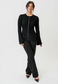 Schwarzer gerippter Cardigan mit langen Ärmeln und geradem Saum, mit einer Knopfleiste auf der Vorderseite, kombiniert mit enganliegenden schwarzen Hosen.