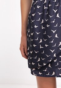 Sininen mekko, jossa on kermansävyinen lintukuosi, pehmeä ja sileä pinta sekä kietaisumalli helmassa, näkyvä käsi pitämässä kangasta.