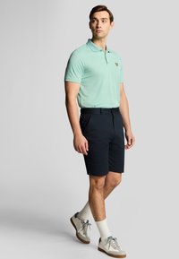 Mintgrøn polo med korte ærmer, navy shorts og lys grå sneakers med blå accenter, stående imod en neutral baggrund.