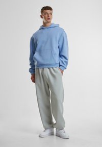 Helles blaues Hoodie mit einer Vorderentasche, kombiniert mit grauen Jogginghosen. Das Model trägt weiße Sneaker und präsentiert ein entspanntes, lässiges Outfit.