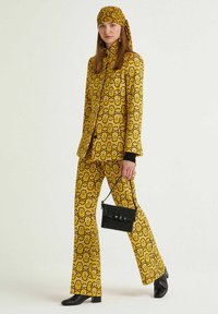 Completo giallo con motivo a disegni geometrici audaci, abbinato a una borsa nera e scarpe con tacco. Tessuto liscio, giacca a maniche lunghe, pantaloni svasati.