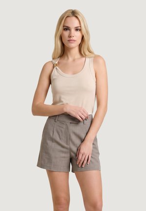 Giovane donna con lunghi capelli biondi indossa un top beige senza maniche con una fibbia e pantaloncini grigi a vita alta, in piedi davanti a uno sfondo uniforme.