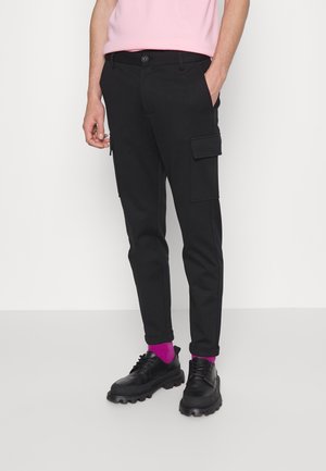 Man draagt zwarte cargobroek met zijzakken, zwarte chunky schoenen, felroze sokken en een lichtroze overhemd, staand tegen een witte achtergrond.