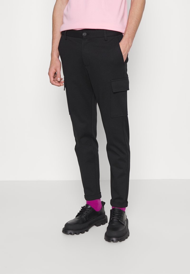 Homme portant un pantalon cargo noir avec des poches latérales, des chaussures grosses noires, des chaussettes rose vif et une chemise rose clair, debout devant un fond blanc.