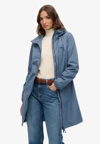 Kobieta w niebieskim płaszczu przeciwdeszczowym z kapturem, na beżowym swetrze z golfem i niebieskich dżinsach z brązowym plecionym paskiem, stojąca z jedną ręką w kieszeni.