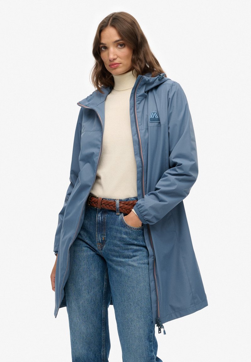 Kobieta w niebieskim płaszczu przeciwdeszczowym z kapturem, na beżowym swetrze z golfem i niebieskich dżinsach z brązowym plecionym paskiem, stojąca z jedną ręką w kieszeni.