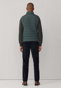 Gewatteerd groen vest met een opstaande kraag, gecombineerd met een donkere trui en zwarte broek. Glad oppervlak en gelijkmatig verdeelde horizontale naden.