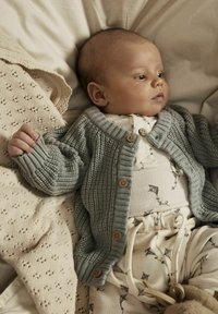 Cardigan lavorato a maglia grigio chiaro con bottoni in legno, abbinato a una tuta crema a fantasia con disegni di aquiloni e pantaloni coordinati, su una superficie morbida.