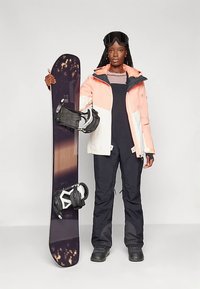 Snowboardzista trzyma czarny snowboard z fioletowymi akcentami, ubrany w koralowo-białą kurtkę, czarne spodnie narciarskie i gogle narciarskie. Widoczne mocowania do snowboardu.