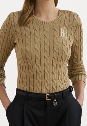 Femme portant un pull torsadé beige avec des boutons sur une épaule et un pantalon noir avec une ceinture noire et un charm en forme de cadenas.