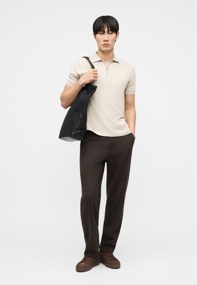 Homme debout portant un polo beige à manches courtes avec fermeture éclair, pantalon foncé, chaussures marron, tenant un sac à bandoulière noir sur un fond clair uni.