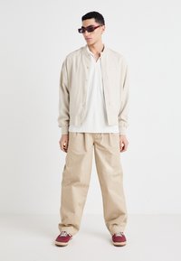 Veste légère beige avec des boutons-pression, portée sur une chemise blanche. Un pantalon beige ample et des baskets bordeaux complètent la tenue.