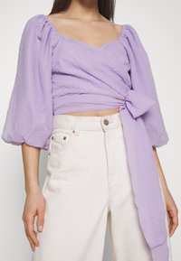 Lavendelwraptop med ballongärmar, texturerad tyg och knytdetalj i sidan, kombinerat med högmidjade vita jeans.