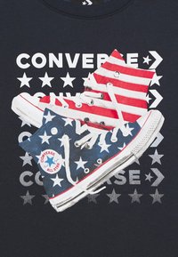 Sneakers Converse Chuck Taylor con strisce rosse e bianche, blu con stelle bianche, esposti su una maglietta nera con testo bianco e grafiche a forma di stella.