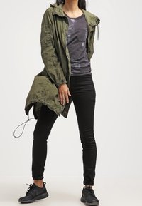 Veste à capuche vert olive sur une chemise grise, assortie à un jean skinny noir et des baskets noires. La veste a une coupe décontractée et un cordon de serrage.