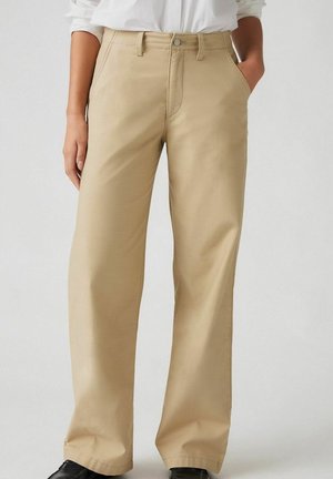 Beige gerade geschnittene Hose, getragen mit einem in die Hose gesteckten weißen Hemd, Hände in den Taschen und schwarzen Schuhen.
