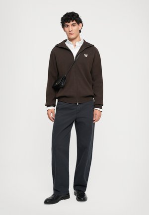Young man wearing a brown zip-up hoodie, white shirt, loose dark pants, black shoes, and a small black crossbody bag against a plain background.