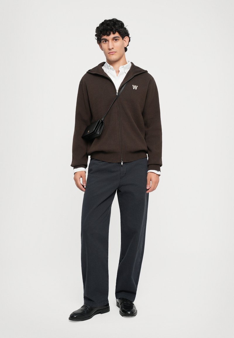Young man wearing a brown zip-up hoodie, white shirt, loose dark pants, black shoes, and a small black crossbody bag against a plain background.