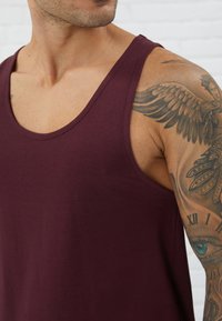 Bordeaux top tank dibuat dari kain lembut dengan leher lebar, desain fit, dan tanpa detail yang terlihat. Menampilkan lengan kekar dengan tato.