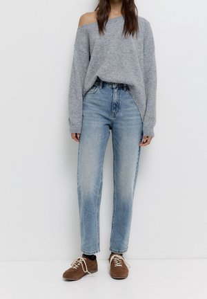 Persoon die een grijze off-shoulder trui, lichtblauwe hoge taille jeans, zwarte sokken en bruine veterschoenen draagt, staand tegen een witte muur.