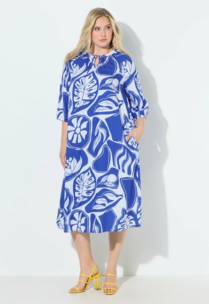 Frau trägt ein blau-weiß geblümtes Midikleid mit ellbogenlangen Ärmeln und gelben Riemchensandaletten mit Absatz, steht an einer weißen Wand.