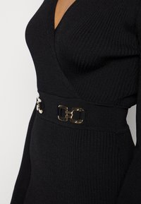 Zwarte geribbelde jurk met een diepe V-hals, voorzien van een decoratieve gouden gespceintuur op de taille voor extra detail. Lange mouwen.