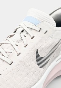 Light gray running shoe with a textured knit upper, black accent logo, and a soft pink sole. Features a blue heel tab and lace closure.