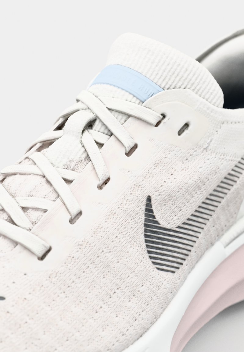 Zapatilla de running gris claro con parte superior de punto texturizado, logotipo de acento negro y suela de un suave color rosa. Presenta un tirador en el talón azul y cierre de cordones.